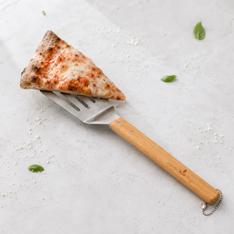A triangular slice of pizza on a metal pizza server with a light wood handle, set on a light gray surface with scattered basil leaves and flour.