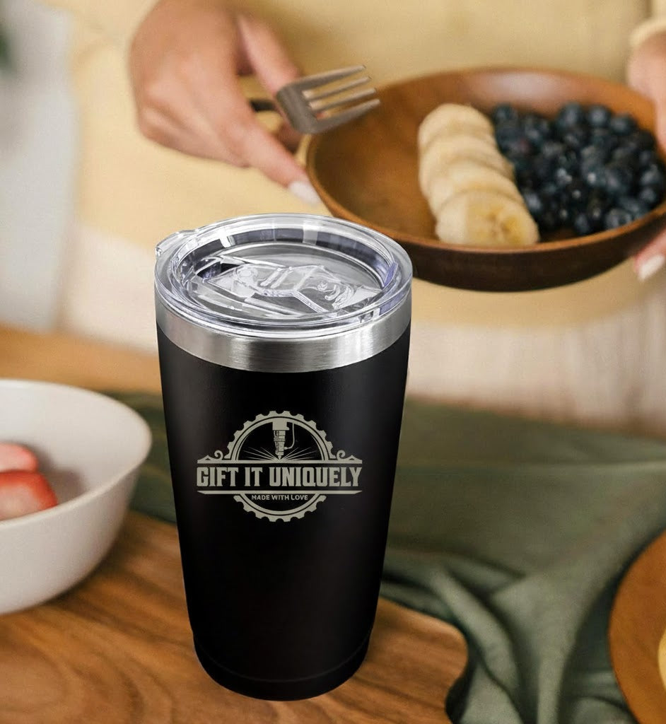 Black tumbler with 'Gift It Uniquely' logo on a wooden table with fruit bowls in the background
