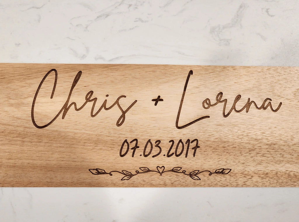 Wooden cutting board with engraved names 'Chris + Lorena' and date '07.03.2017' on a marble background