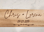 Wooden cutting board with engraved names 'Chris + Lorena' and date '07.03.2017' on a marble background
