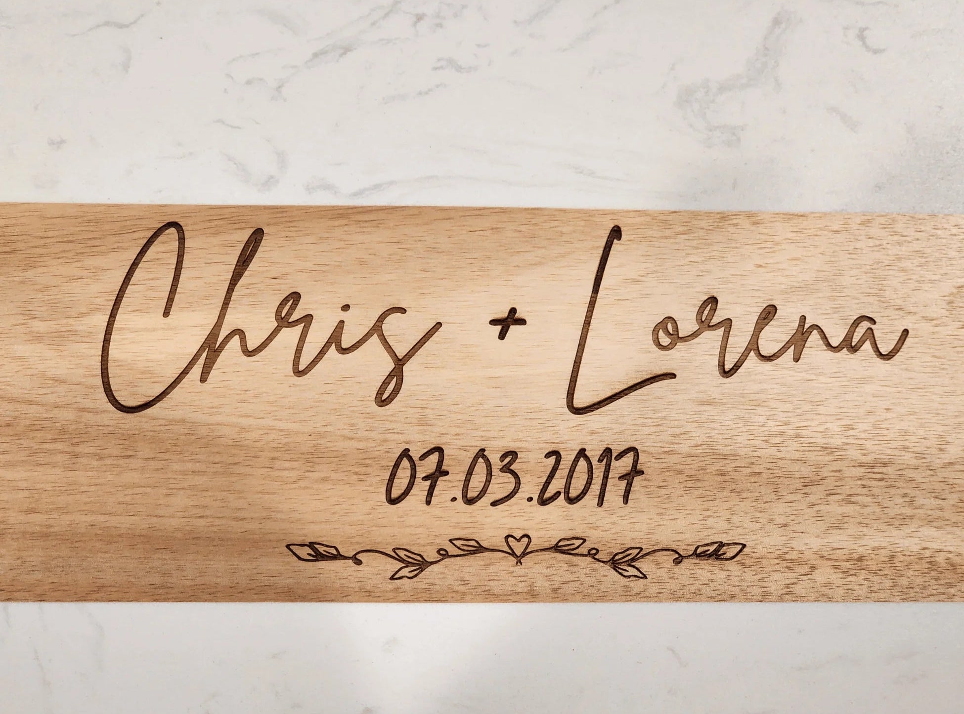 Wooden cutting board with engraved names 'Chris + Lorena' and date '07.03.2017' on a marble background