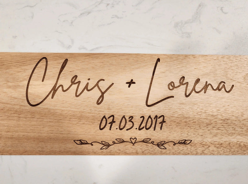 Wooden cutting board with engraved names 'Chris + Lorena' and date '07.03.2017' on a marble background
