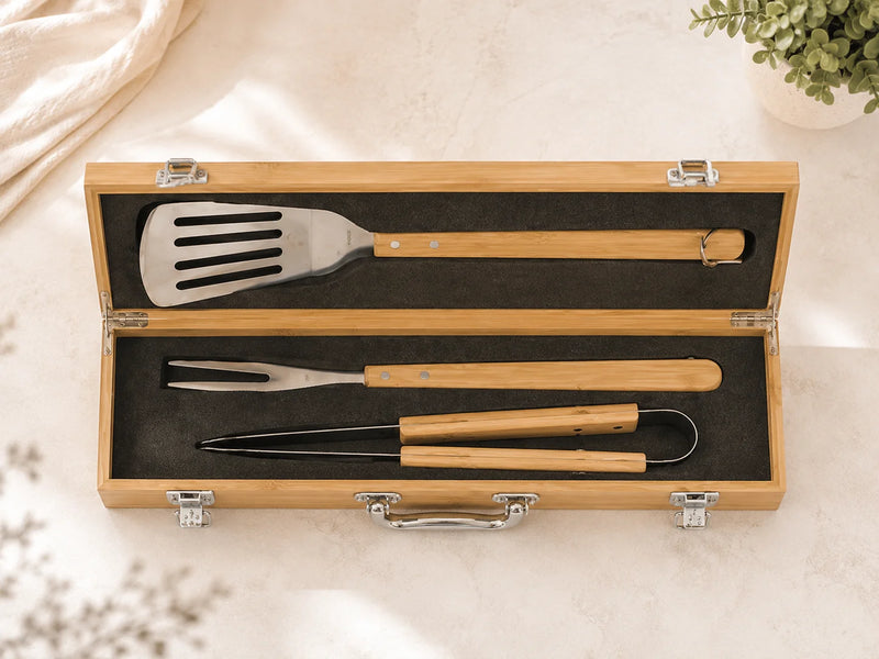 A 3-piece barbecue tool set with stainless steel heads and light wood handles, neatly arranged inside an open light wood carrying case with a black foam insert, photographed on a light beige stone surface.