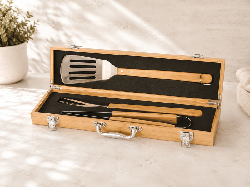 A 3-piece barbecue tool set with wooden handles and stainless steel heads, presented in an open bamboo carrying case on a light stone surface.