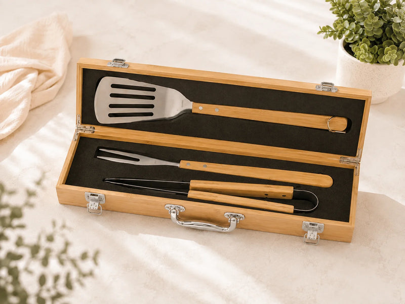 A 3-piece outdoor BBQ tool set with bamboo handles and stainless steel heads, presented in an open light wood carrying case on a cream fabric surface.