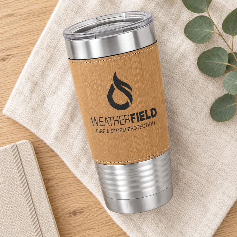 A Weatherfield insulated tumbler with a natural wood-textured wrap and stainless steel accents, shown on a beige cloth over a wooden surface with eucalyptus leaves and a notebook nearby.