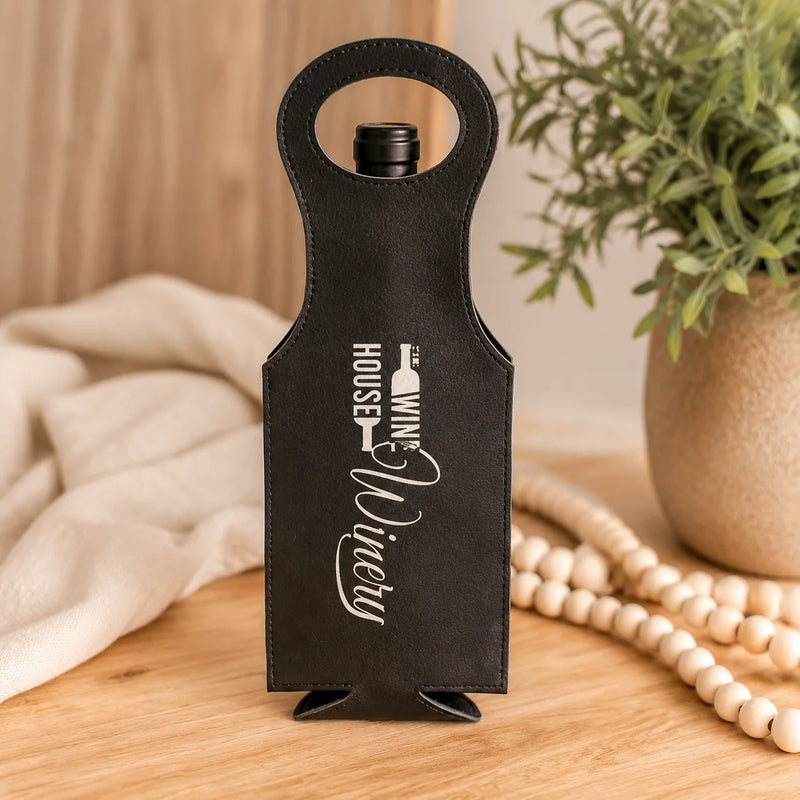 A black faux-leather wine bottle carrier with a loop handle and white 'Wine Nicely' branding, standing on a wooden table with a beige cloth, wooden beads, and a potted plant in the background.