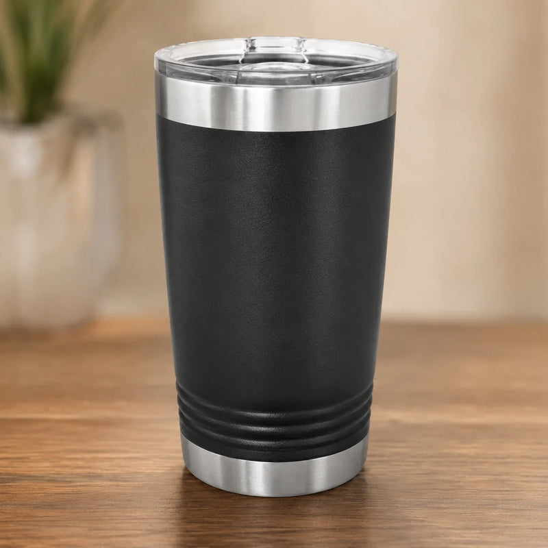 A black insulated travel tumbler with a clear lid and brushed stainless steel trim, standing on a wooden tabletop with a softly blurred indoor background.