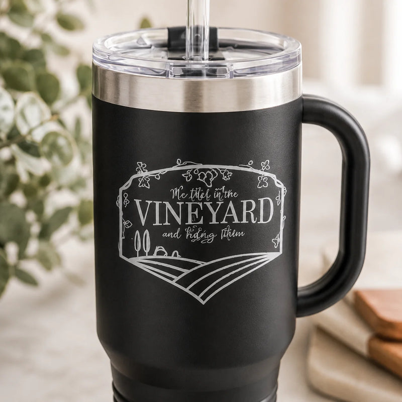 A black insulated travel tumbler with a clear lid and straw, featuring a white engraved vineyard-themed design on the front. The tumbler is shown in a softly blurred indoor setting with greenery and neutral surfaces in the background.