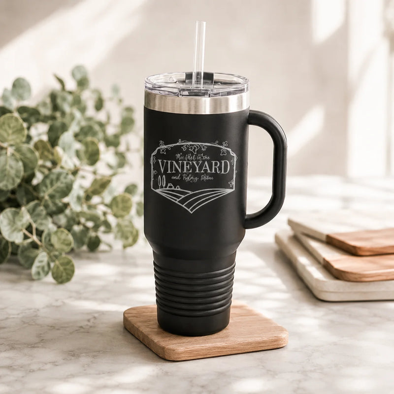 A black insulated travel tumbler with a handle, clear lid, and straw, standing on a wooden coaster on a marble surface. The tumbler is engraved with the words 'The Best of the VINEYARD and Vine Bar.