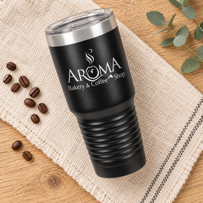 A black insulated tumbler with a clear lid sits on a beige woven cloth over a wooden surface, with coffee beans and eucalyptus leaves around it. The tumbler is printed with the text 'AROMA Bakery & Coffee Shop'.