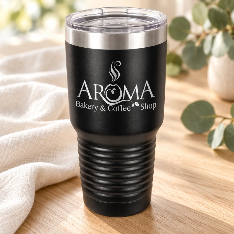 A black insulated tumbler with a clear lid sits on a wooden surface next to a cream cloth and green leaves. The tumbler is printed with the text 'AROMA Bakery & Coffee Shop' in white.