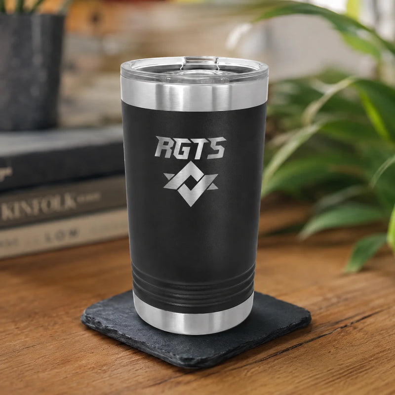 A black insulated tumbler with silver stainless steel trim and a clear lid, standing on a dark slate coaster on a wooden table. The tumbler is engraved with the text 'RGTS' and a geometric logo.