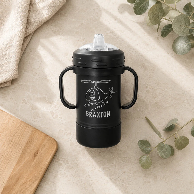A black insulated tumbler with two side handles and a clear lid, featuring a white helicopter illustration and the name BRAXTON, styled on a beige stone surface with a cloth, wood board, and eucalyptus leaves.