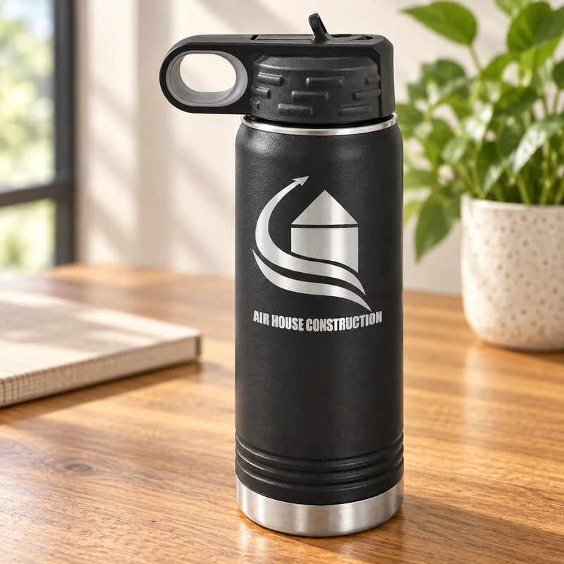 A black insulated water bottle with a silver and white 'Air House Construction' logo, standing on a wooden table with a blurred background of a window, notebook, and potted plant.