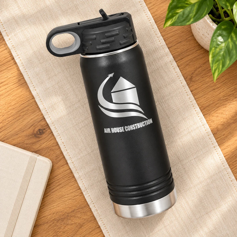 A black insulated water bottle with a silver logo and 'AIR HOUSE CONSTRUCTION' text, placed diagonally on a beige fabric runner over a wooden surface with a plant leaf in the corner.