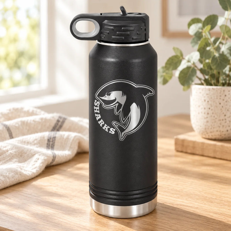 A black insulated water bottle with a silver shark graphic and the word 'SHARKS' on the front, shown standing on a wooden table in a bright indoor setting with a plant and window in the background.