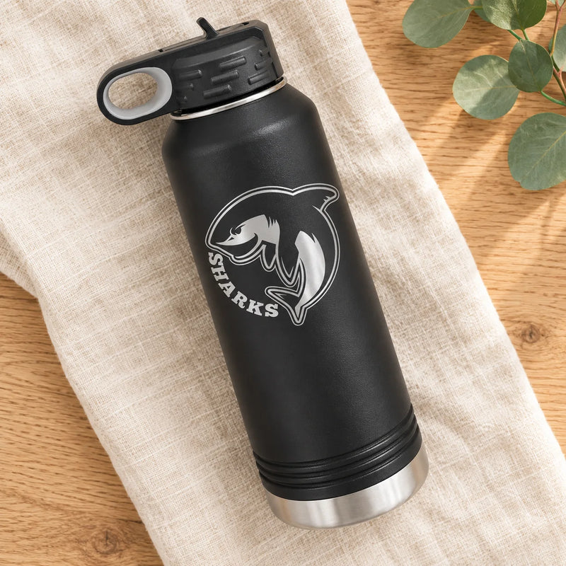 A black insulated water bottle with a silver shark logo and the word 'SHARKS' on the front, resting on a beige linen cloth over a wooden surface with green leaves in the corner.