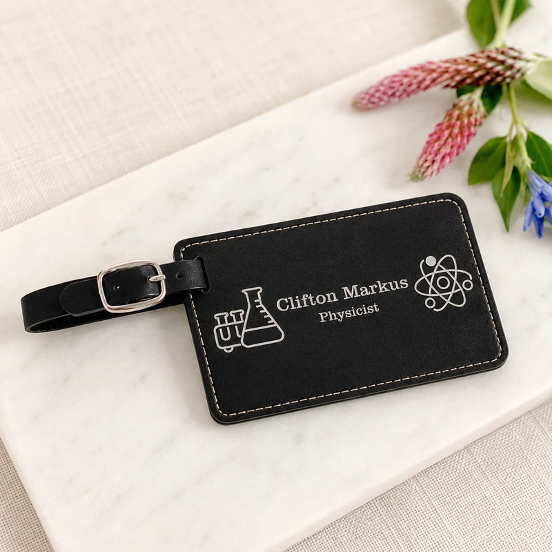 A black leather luggage tag with a silver buckle, featuring white engraved text reading 'Clifton Markus Physicist' along with science icons, styled on a white marble block with decorative flowers.