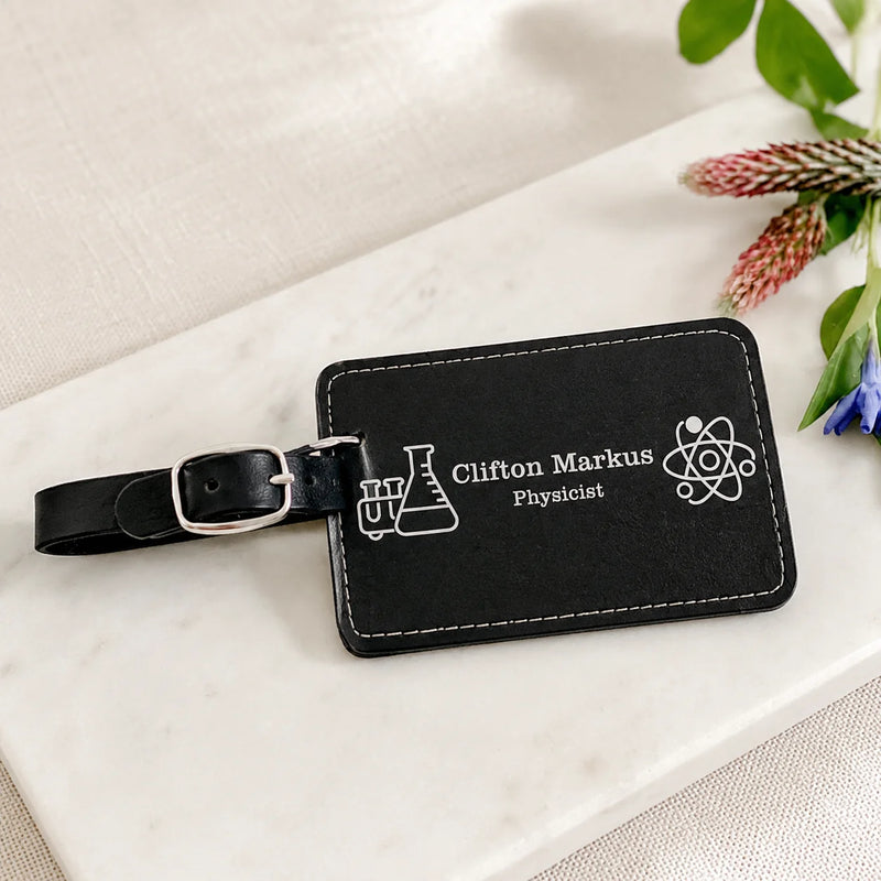 A black leather luggage tag with a silver buckle strap, featuring white printed text reading 'Clifton Markus Physicist' along with science-themed icons, displayed on a white marble surface with decorative flowers.