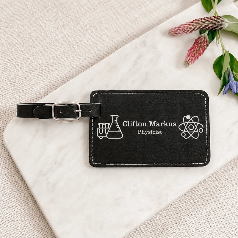 A black personalized luggage tag with white engraved text reading 'Clifton Markus Physicist' and science-themed icons, shown on a white marble board with fabric and small flowers around it.
