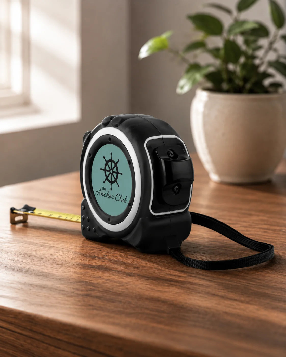 A black retractable tape measure with a mint green circular face and a ship's wheel logo, resting on a wooden table with its yellow tape extended. A potted plant is visible in the background.