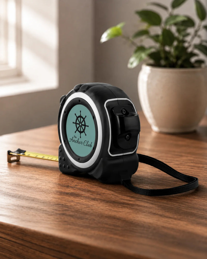A black retractable tape measure with a mint green circular face and a ship's wheel logo, resting on a wooden table with its yellow tape extended. A potted plant is visible in the background.