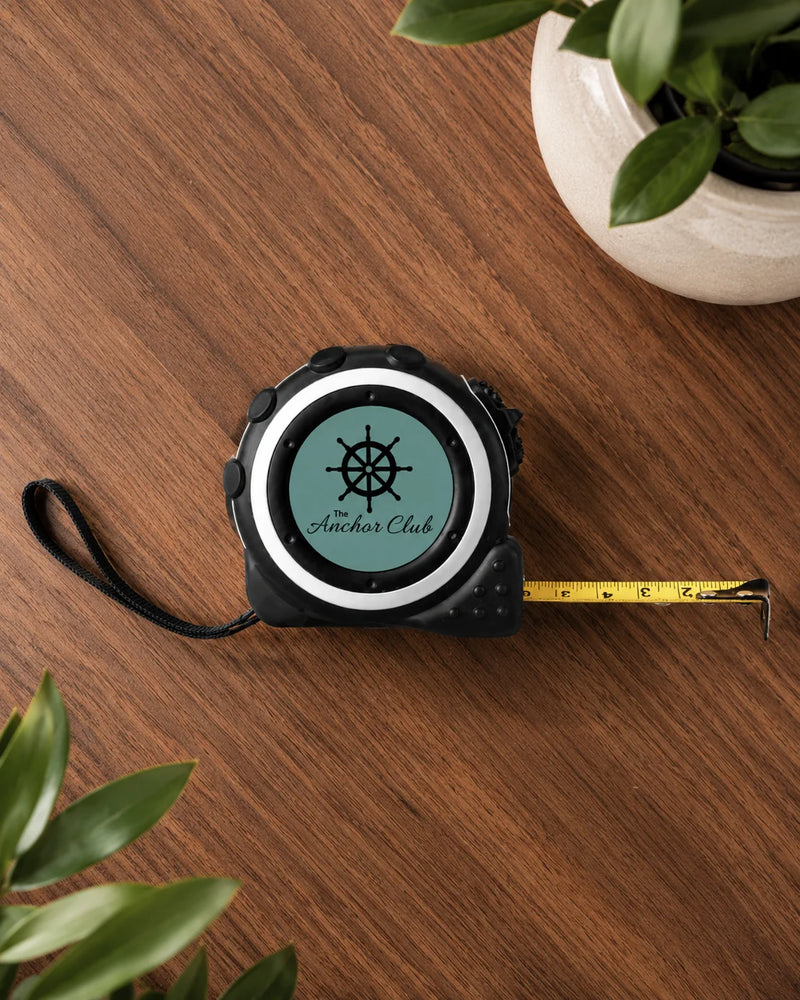 A black tape measure with a yellow retractable scale lies on a wooden tabletop. The tape measure face is personalized with a circular teal insert featuring a black nautical wheel logo and the text 'The Anchor Club'.