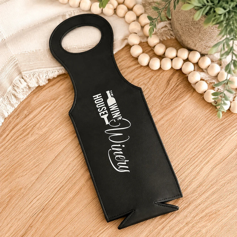 A black wine bottle opener with white printed text reading 'The Wine House Vinicius' resting on a light wood tabletop, styled with a cream fringed cloth, wooden beads, and a small plant.