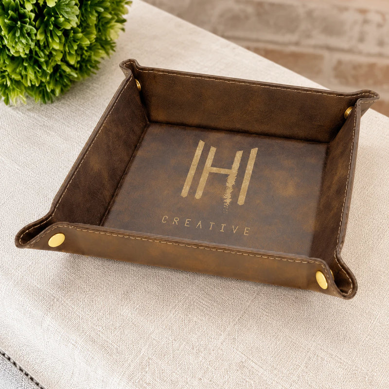 A brown faux-leather desk organizer tray with gold hardware and a gold-foiled 'IH Creative' logo on the base, shown on a textured beige tablecloth with a green plant in the background.