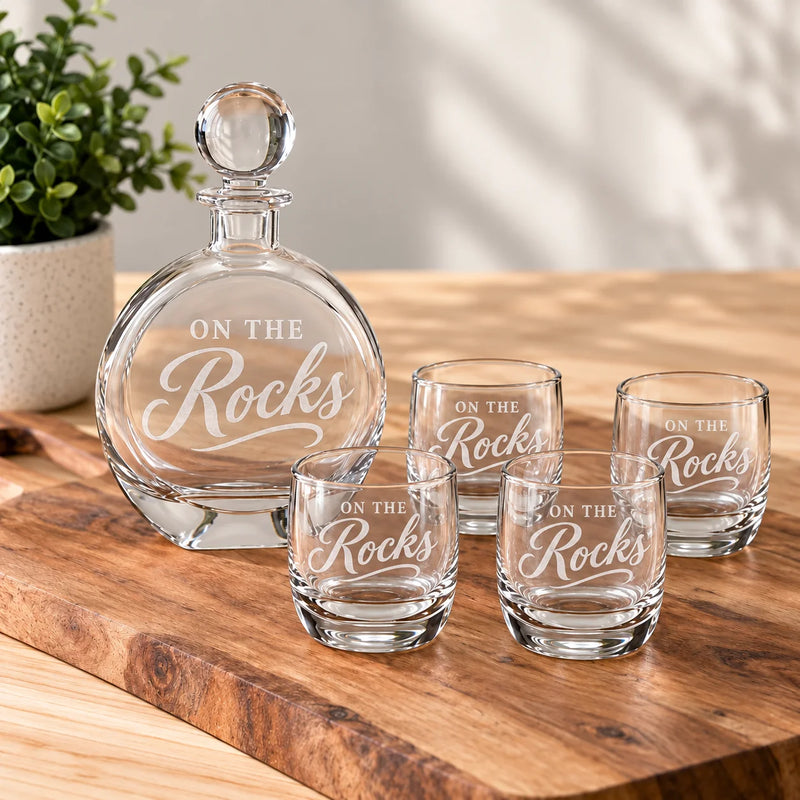 A clear glass decanter and four matching rocks glasses engraved with the phrase 'ON THE Rocks', arranged on a wooden serving board with a potted plant in the background.