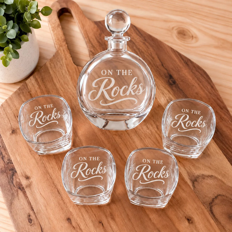 A clear glass decanter and four matching shot glasses engraved with the phrase 'ON THE Rocks,' arranged on a wooden serving board with a small potted plant in the background.