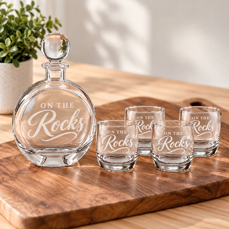 A clear glass decanter with a matching stopper and four matching shot glasses, all etched with the phrase 'ON THE Rocks,' arranged on a wooden serving board in a bright indoor setting with a potted plant in the background.
