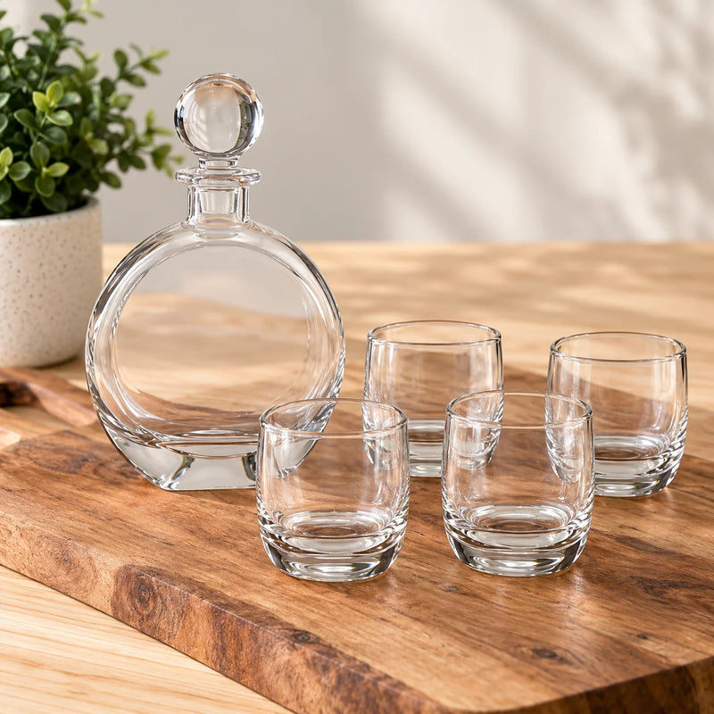 A clear glass decanter with a round stopper and four matching small clear glasses arranged on a wooden board, with a potted plant in the background.