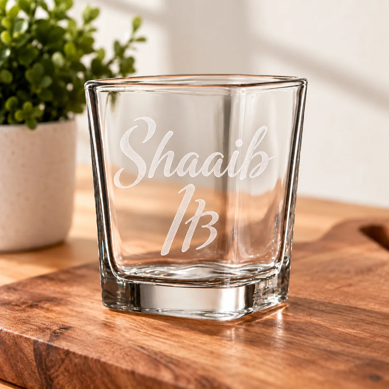 A clear glass rocks tumbler with the name 'Shaaib' and number '13' etched on the front, sitting on a wooden cutting board with a potted plant in the background.