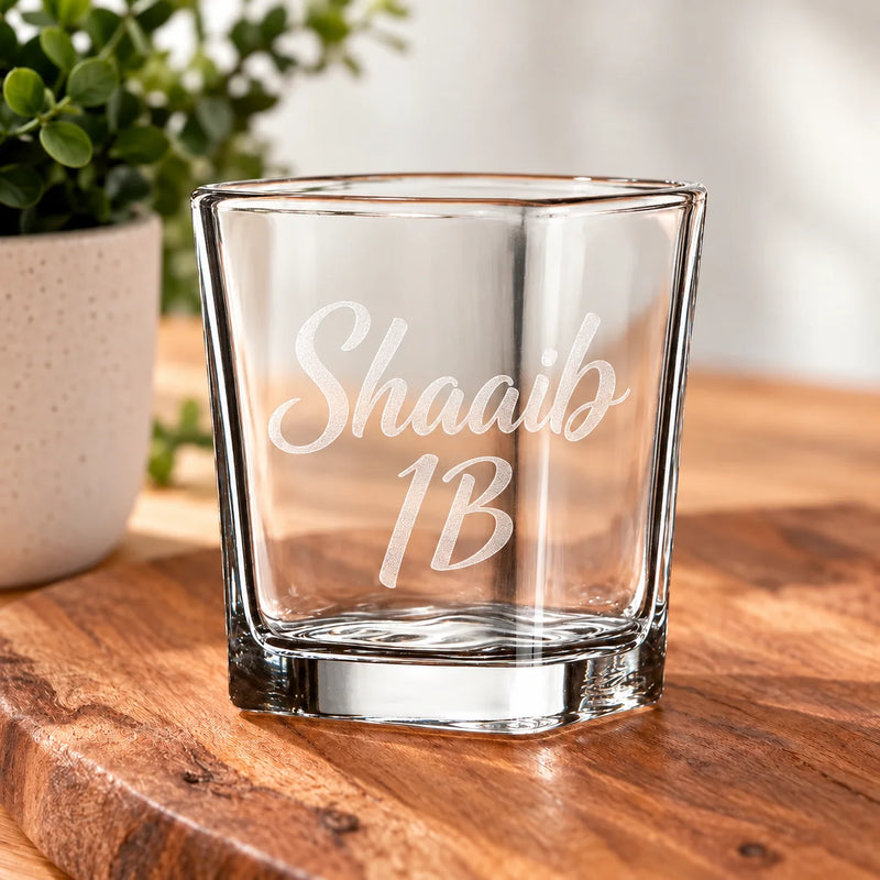 A clear glass rocks tumbler with the personalized name 'Shaaib 1B' etched on the front, sitting on a wooden surface with a blurred potted plant in the background.