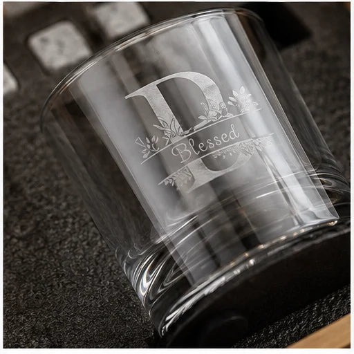 A clear glass tumbler with a custom monogram 'D' and the word 'Blessed' etched on the front, resting on a dark textured surface.