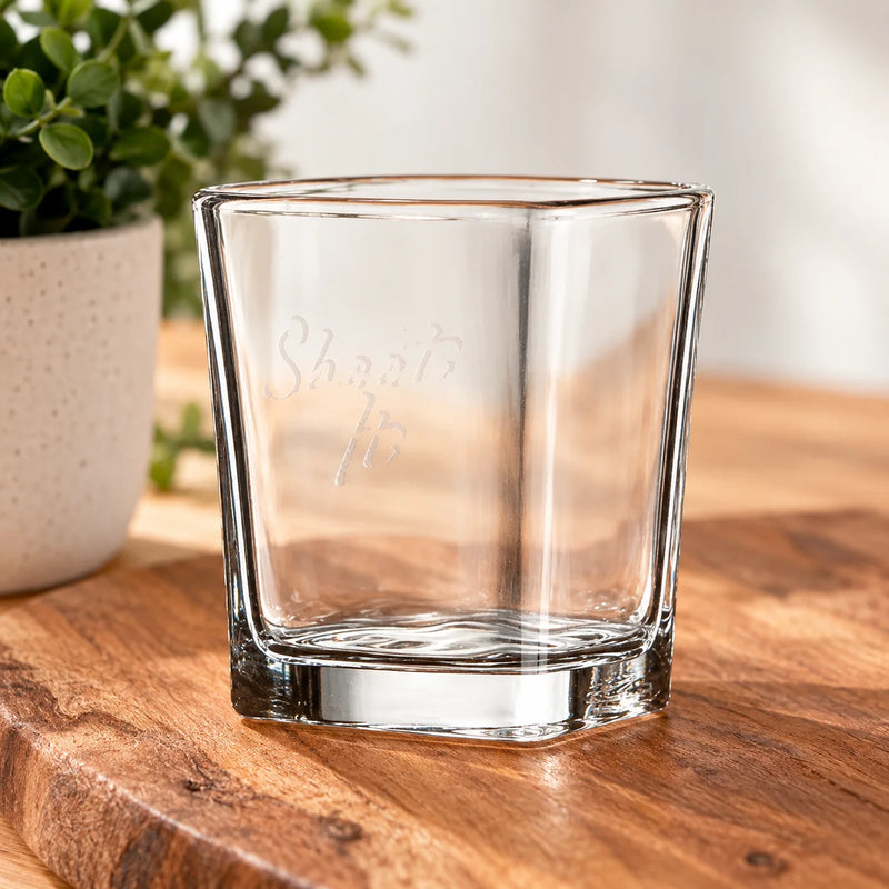 A clear octagonal glass tumbler with 'Shots to' etched on the front, resting on a wooden cutting board with a blurred potted plant in the background.