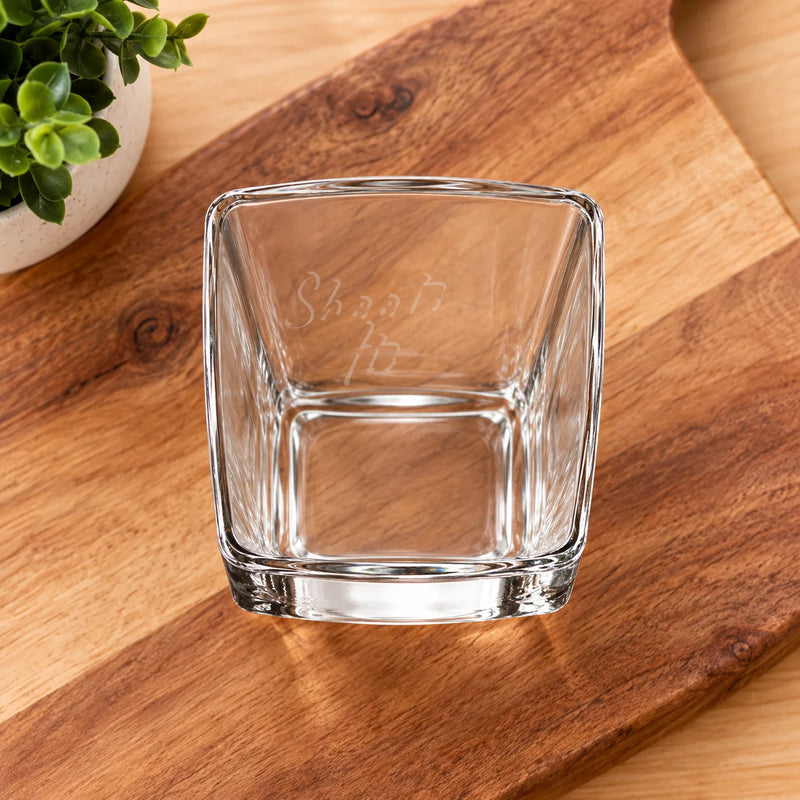 A clear square glass tumbler with a thick base sits on a wooden cutting board, with a small potted green plant partially visible in the top-left corner. The glass has faint etched text inside.