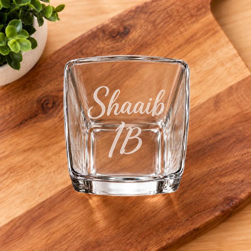 A clear square glass tumbler with the words 'ShaaiB 1B' etched on the front, placed on a wooden cutting board with a small potted plant in the corner.