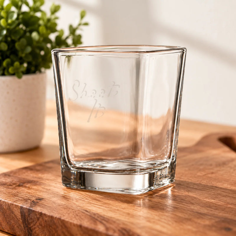 A clear square rocks glass with a thick base, featuring the words 'Shea'sibs' etched on the front, resting on a wooden cutting board with a potted plant in the background.