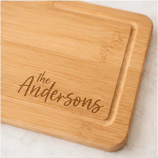 A close-up of a rectangular bamboo cutting board with rounded corners and engraved text reading 'the Andersons' in a dark script, placed on a light stone surface.