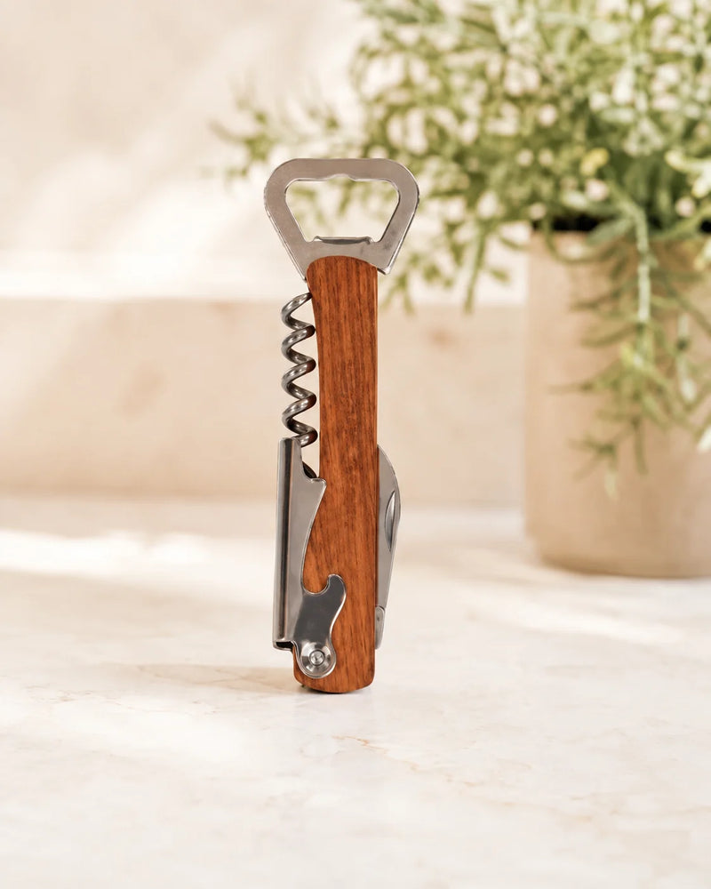 A corkscrew bottle opener with a warm brown wood handle and silver metal tools, standing upright on a light beige surface with a blurred potted plant in the background.