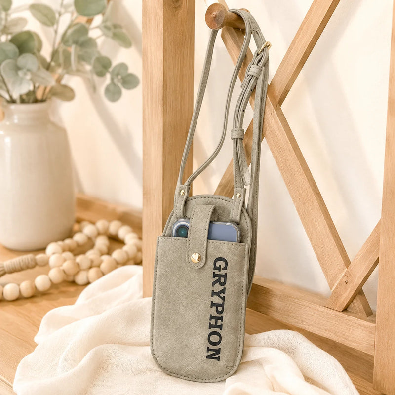 A gray faux-leather crossbody phone pouch with 'GRAYPHON' printed vertically in black, hanging on a wooden chair in a styled indoor setting.