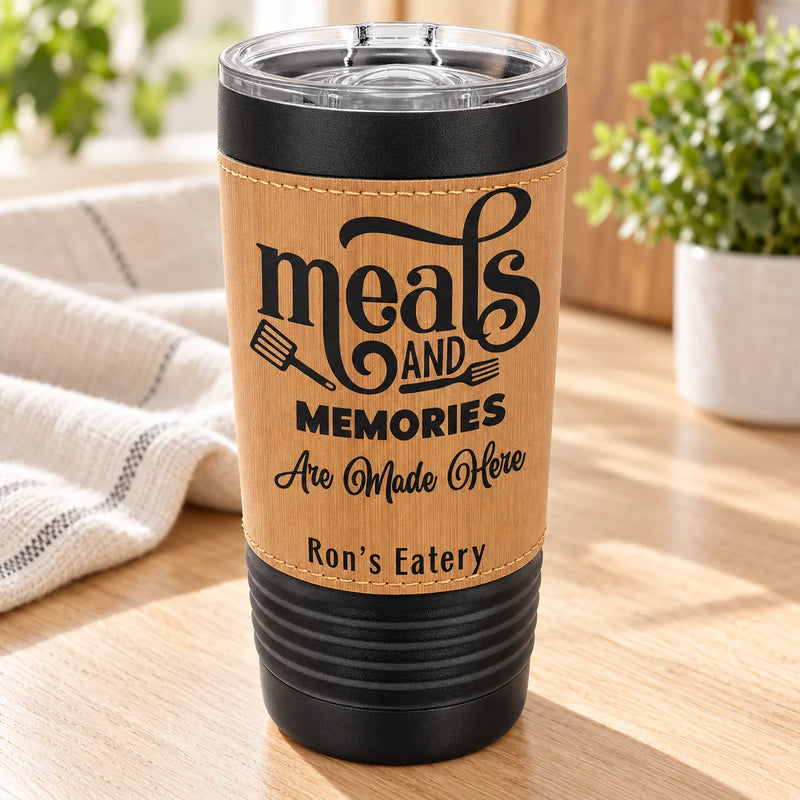 A insulated tumbler with a clear lid sits on a wooden table in a kitchen setting. The tumbler has a light woodgrain-style wrap with black text reading “meals and memories Are Made Here” and “Ron’s Eatery,” plus a small spatula graphic.