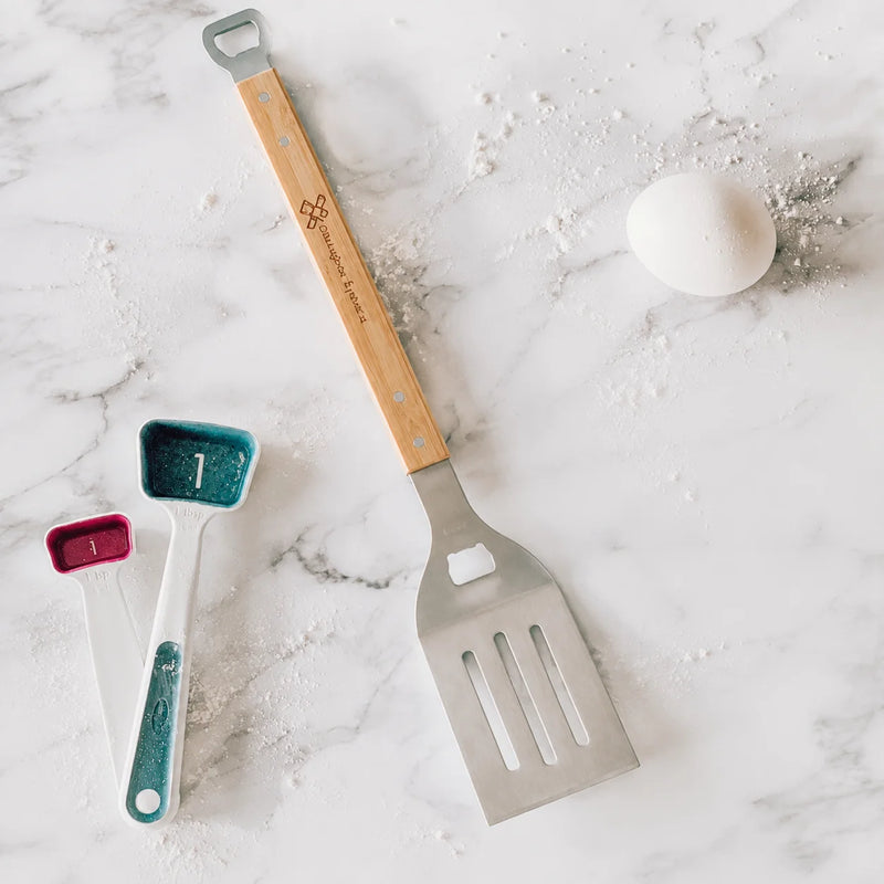 A kitchen utensil set arranged on a white marble surface with flour scattered around. The set includes a large metal spatula with a light wood handle, two smaller measuring spoons in teal and pink, and a white egg prop.