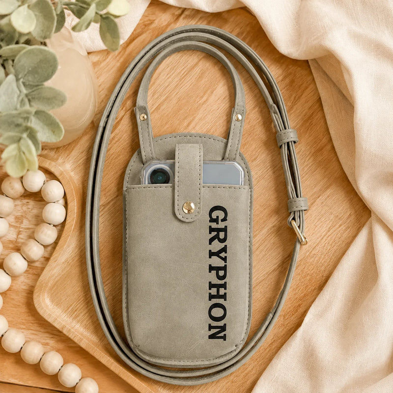 A light gray suede phone pouch with a matching shoulder strap, featuring the word 'GRYPHON' printed vertically in black. The pouch is styled in a flat-lay arrangement on a wooden tray with neutral fabrics, beads, and greenery.