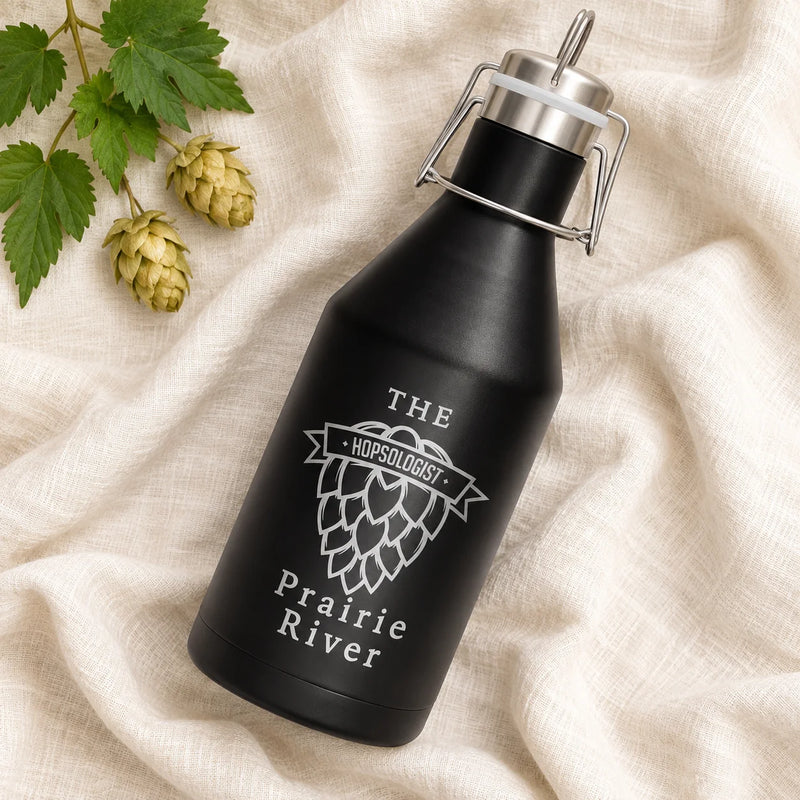 A matte black insulated growler bottle with a metal swing-top lid, placed on draped cream fabric with hops branches and cones as styling props. The bottle is printed with white text reading “THE HOPSLOGIST” and “Prairie River.