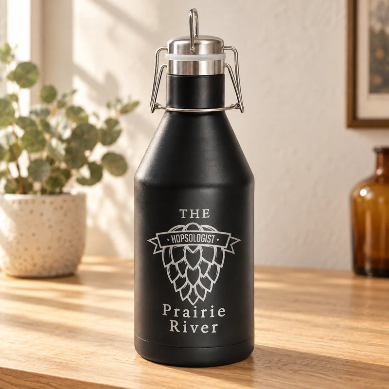 A matte black insulated growler bottle with a swing-top lid, standing on a wooden table in a warm indoor setting. The bottle is printed with a white hop cone graphic and the text 'THE HOPSLOGISTY Prairie River'.