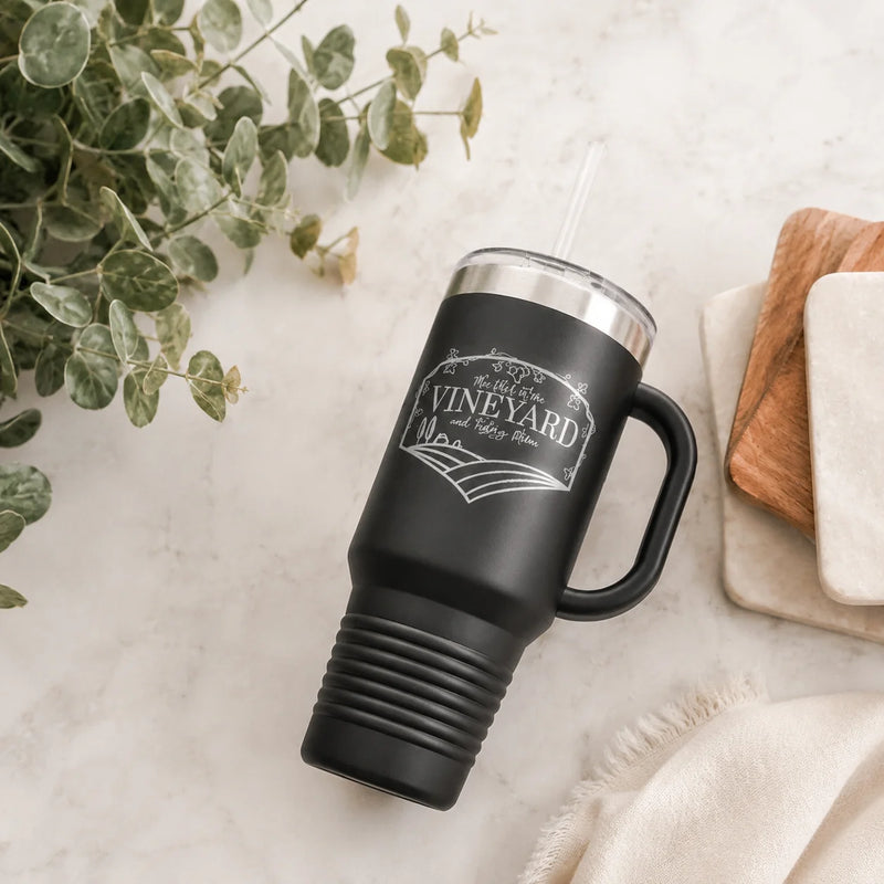 A matte black insulated tumbler with a handle, clear lid, and straw, featuring a white engraved design that reads 'Sip from the VINEYARD and taste them.' It is photographed on a light marble surface with eucalyptus leaves, a wooden board, and a folded cream cloth nearby.